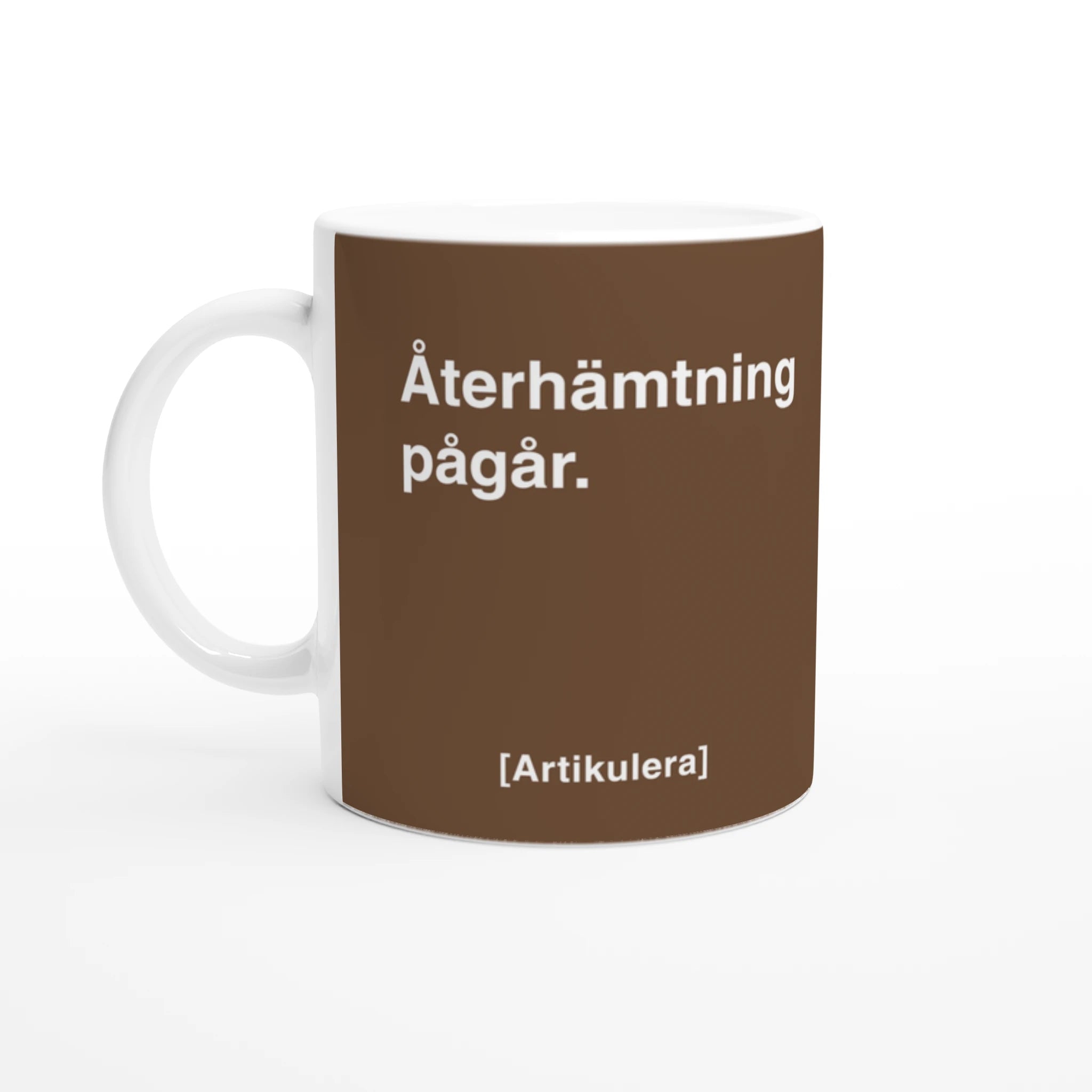 Vit mugg med brunt tryck, vit text ”Återhämtning pågår.” samt vit Artikulera-logotyp