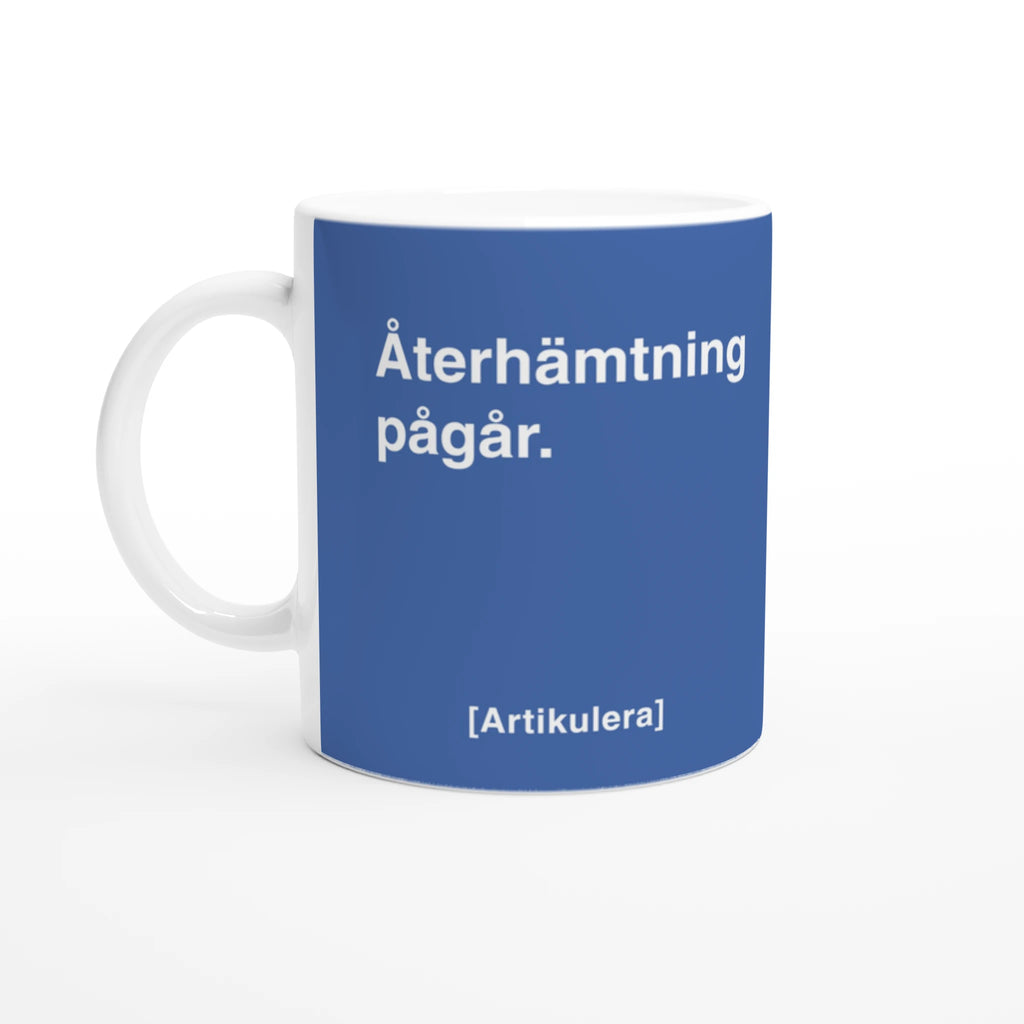 Vit mugg med duvblått tryck, vit text ”Återhämtning pågår.” samt vit Artikulera-logotyp