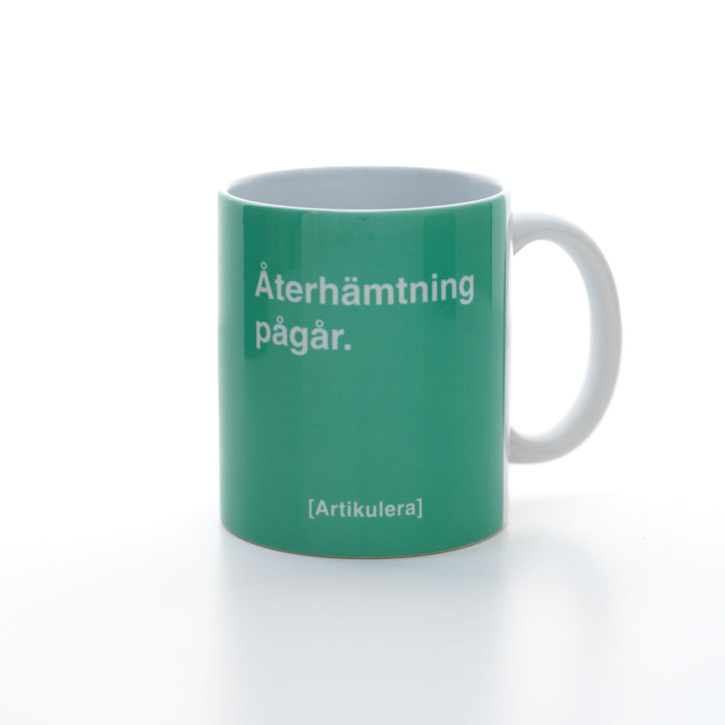 Vit mugg med grönt tryck, vit text ”Återhämtning pågår.” samt vit Artikulera-logotyp