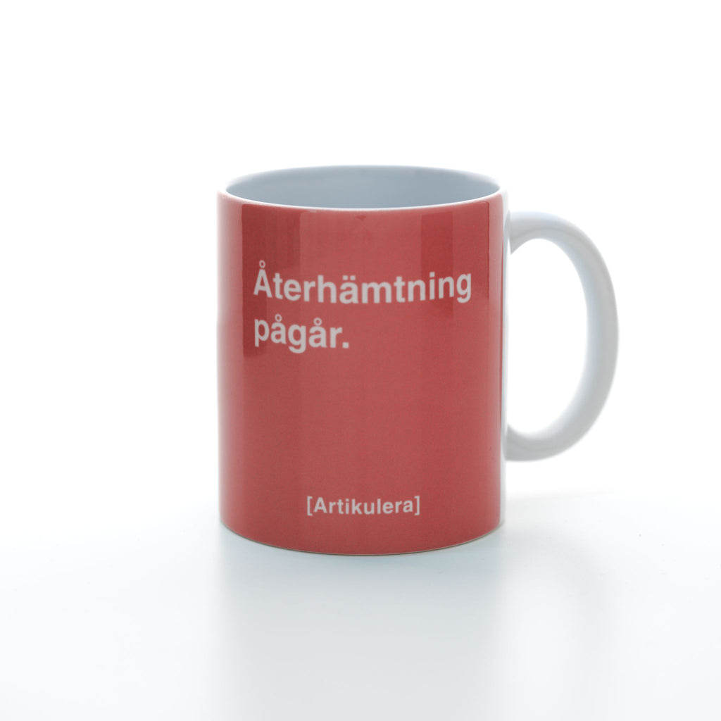 Vit mugg med korallrött tryck, vit text ”Återhämtning pågår.” samt vit Artikulera-logotyp
