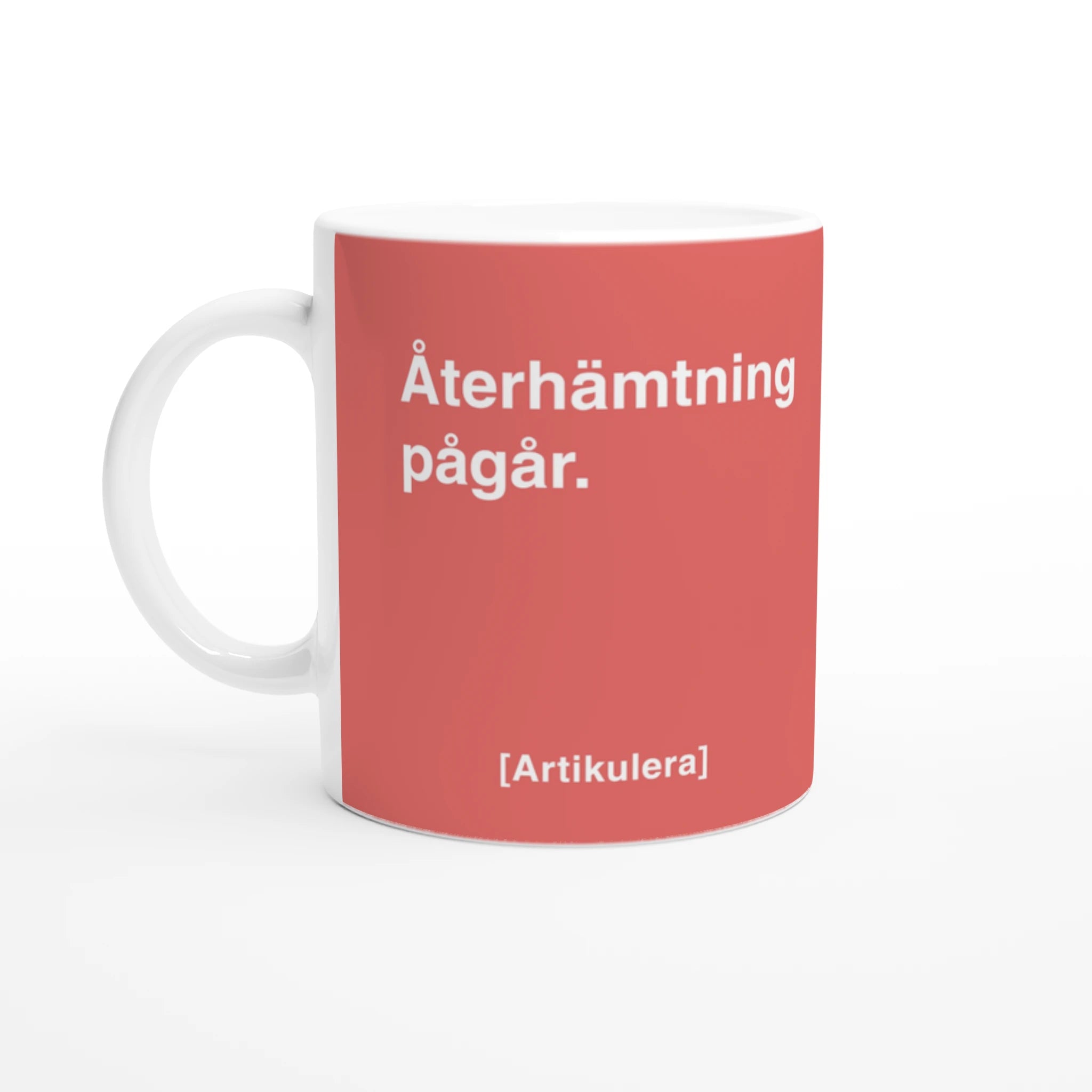 Vit mugg med korallrött tryck, vit text ”Återhämtning pågår.” samt vit Artikulera-logotyp