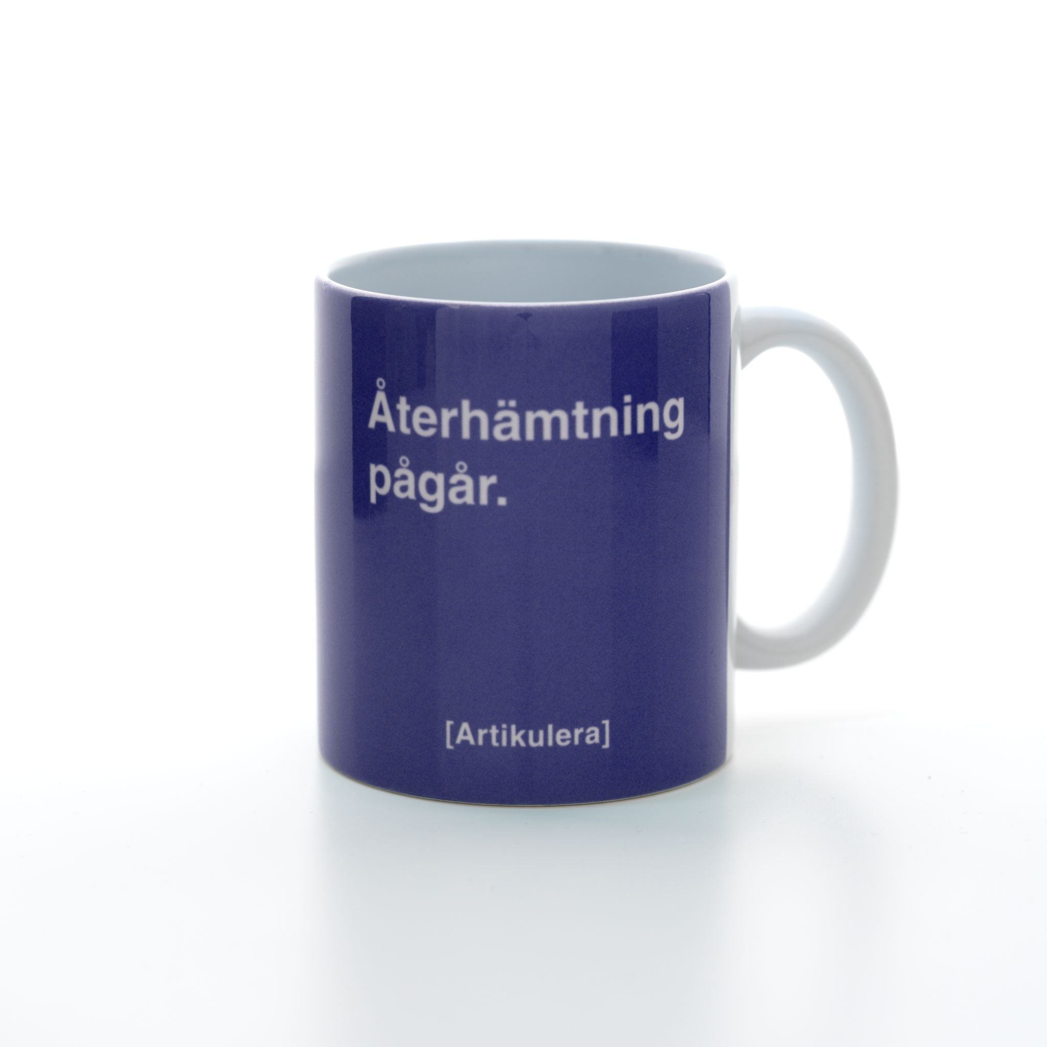Vit mugg med lila tryck, vit text ”Återhämtning pågår.” samt vit Artikulera-logotyp