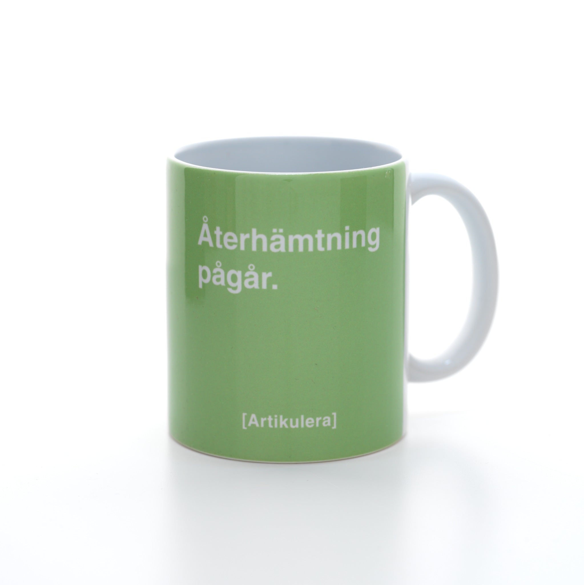 Vit mugg med ljus limegrönt tryck, vit text ”Återhämtning pågår.” samt vit Artikulera-logotyp