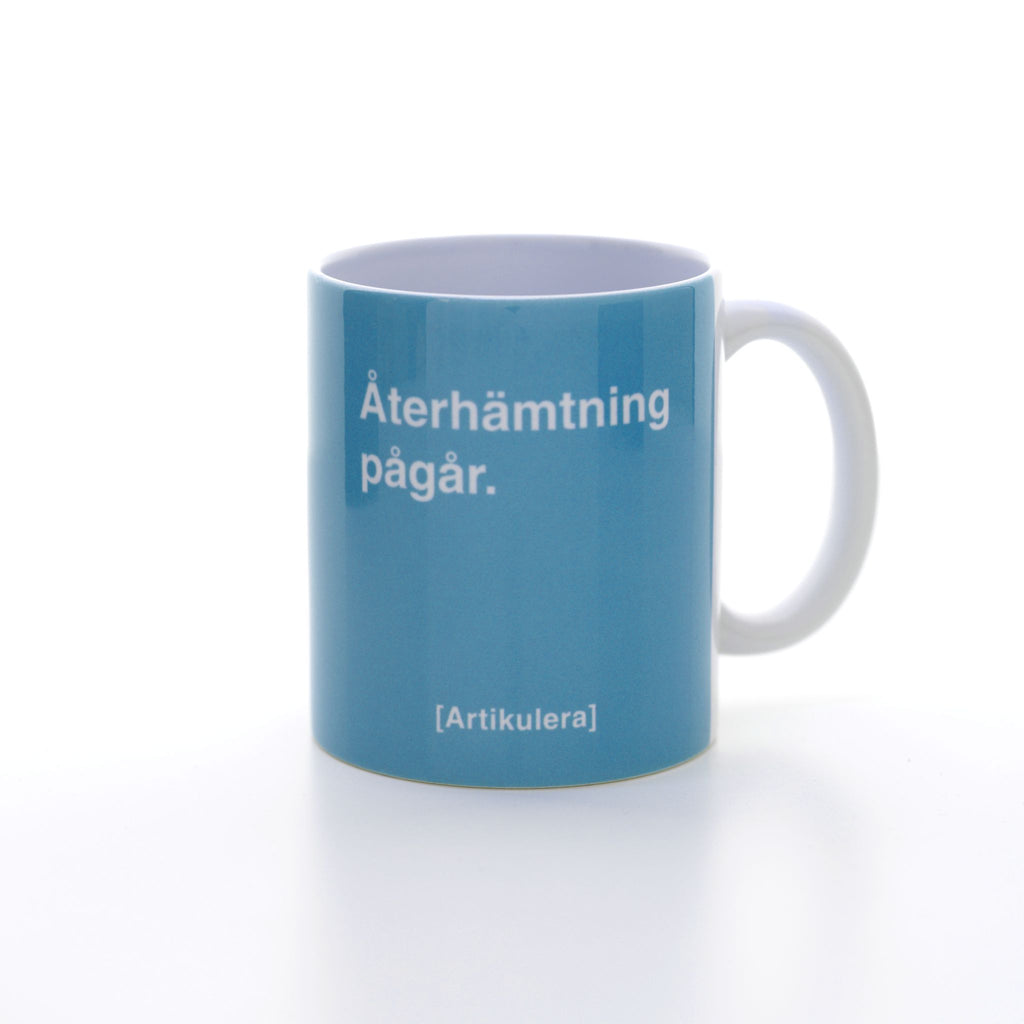 Vit mugg med ljusblått tryck, vit text ”Återhämtning pågår.” samt vit Artikulera-logotyp