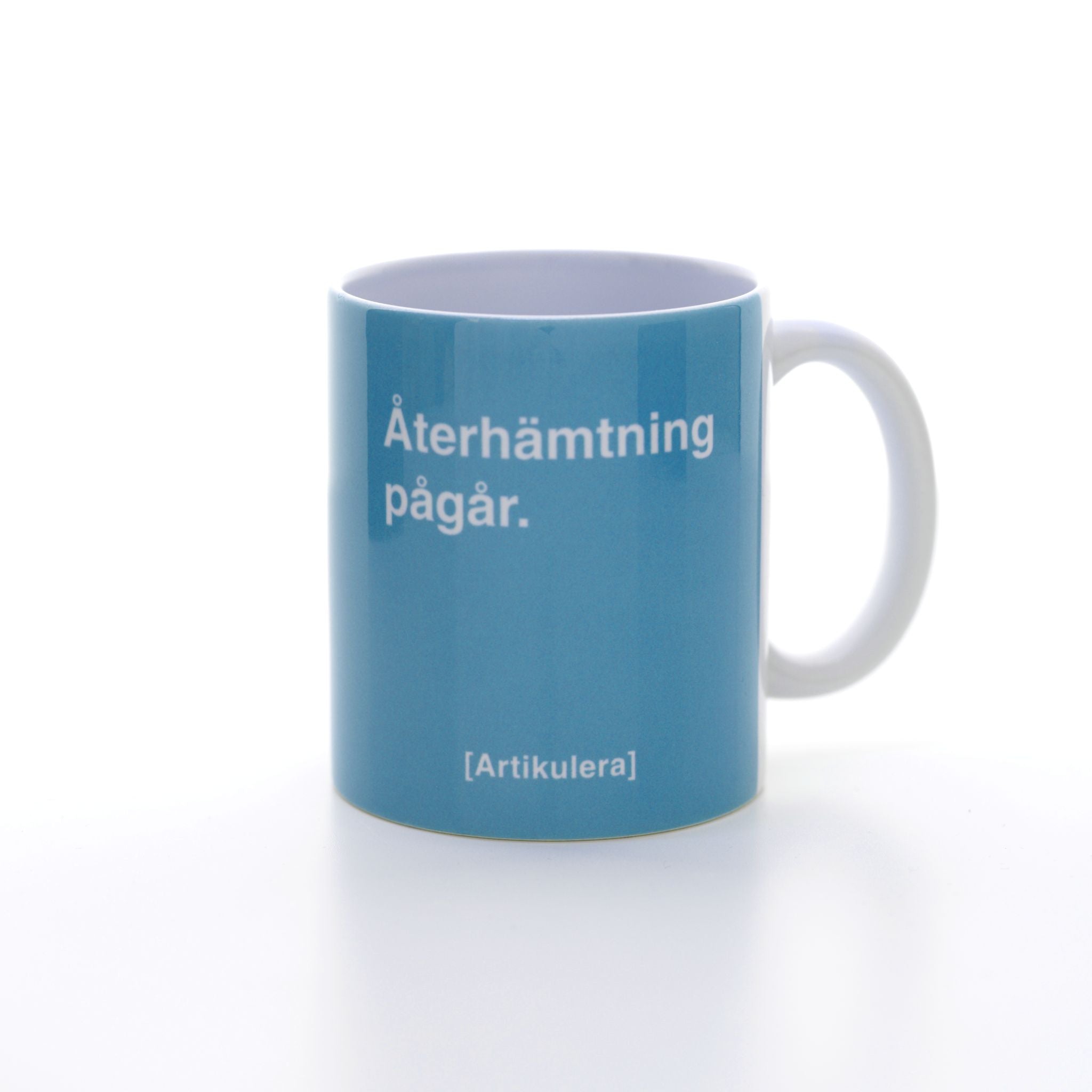 Vit mugg med ljusblått tryck, vit text ”Återhämtning pågår.” samt vit Artikulera-logotyp