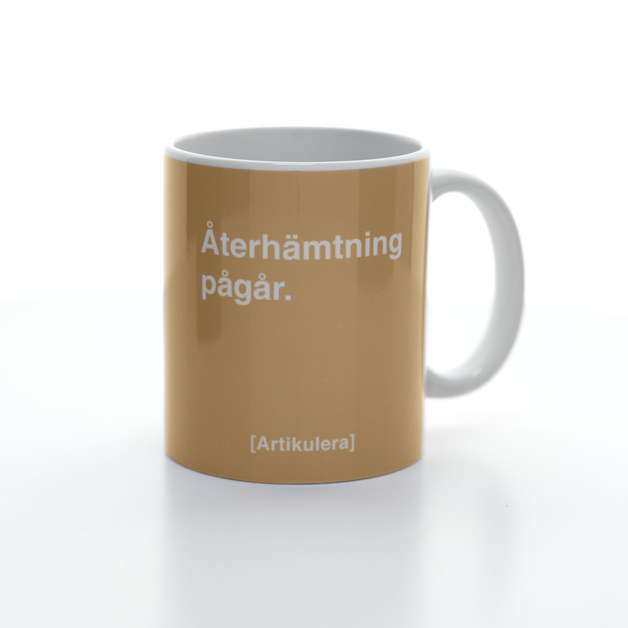 Vit mugg med ljusbrunt tryck, vit text ”Återhämtning pågår.” samt vit Artikulera-logotyp