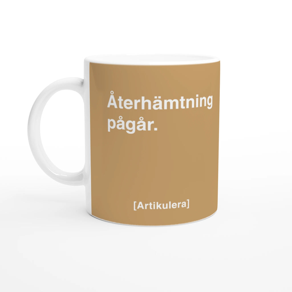 Vit mugg med ljusbrunt tryck, vit text ”Återhämtning pågår.” samt vit Artikulera-logotyp