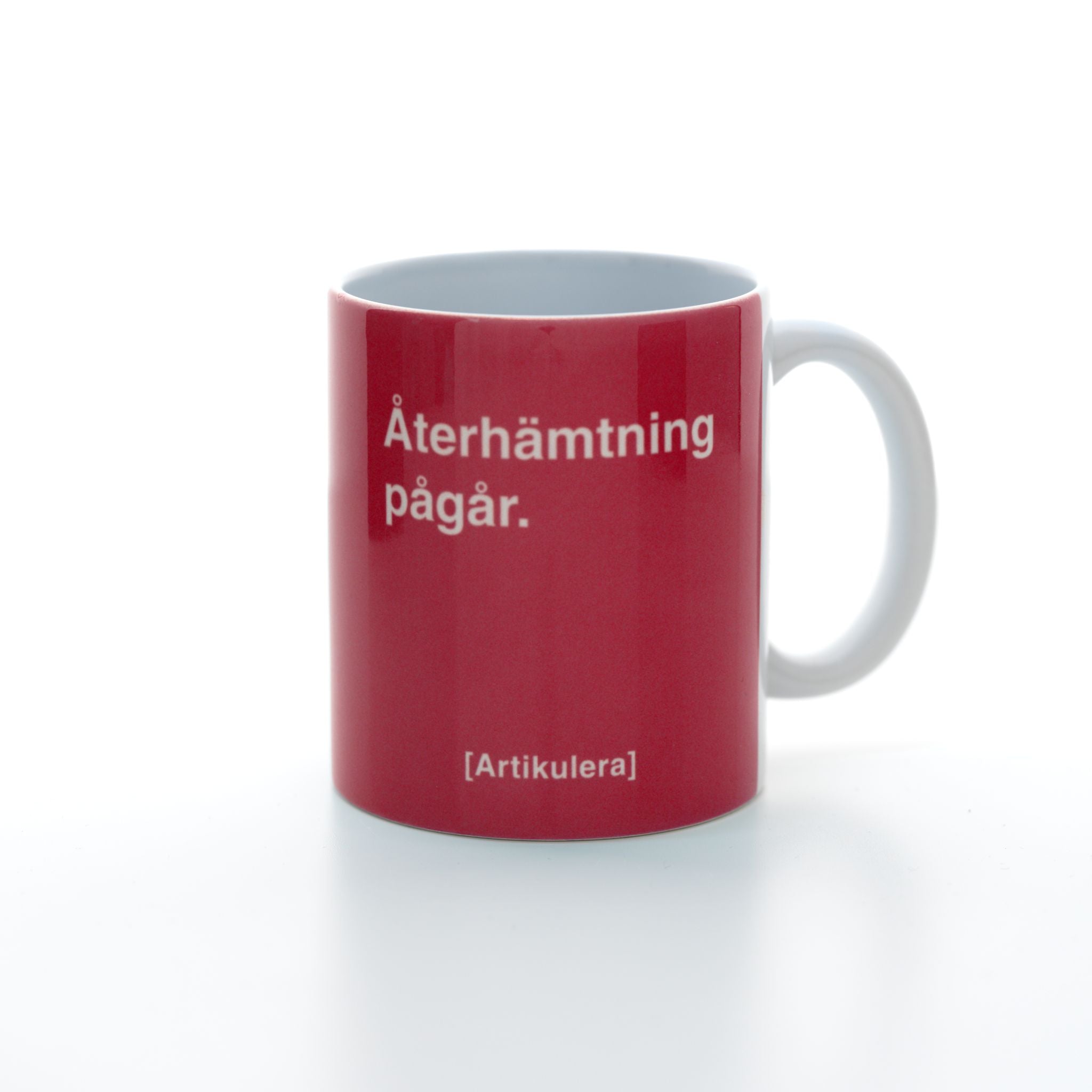 Vit mugg med magentarött tryck, vit text ”Återhämtning pågår.” samt vit Artikulera-logotyp