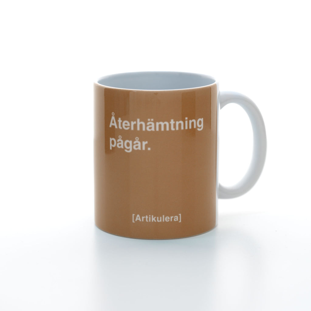 Vit mugg med mediumbrunt tryck, vit text ”Återhämtning pågår.” samt vit Artikulera-logotyp