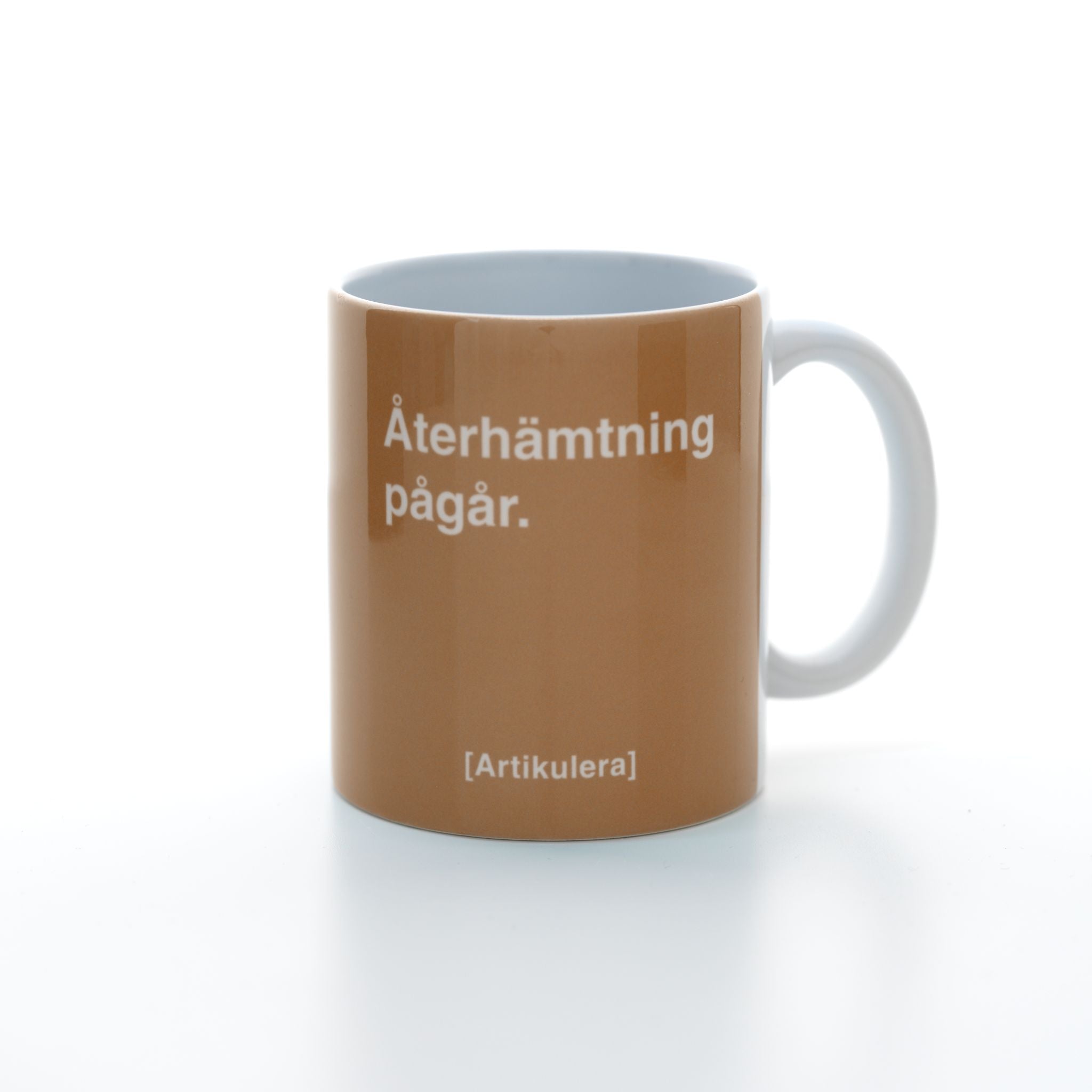 Vit mugg med mediumbrunt tryck, vit text ”Återhämtning pågår.” samt vit Artikulera-logotyp