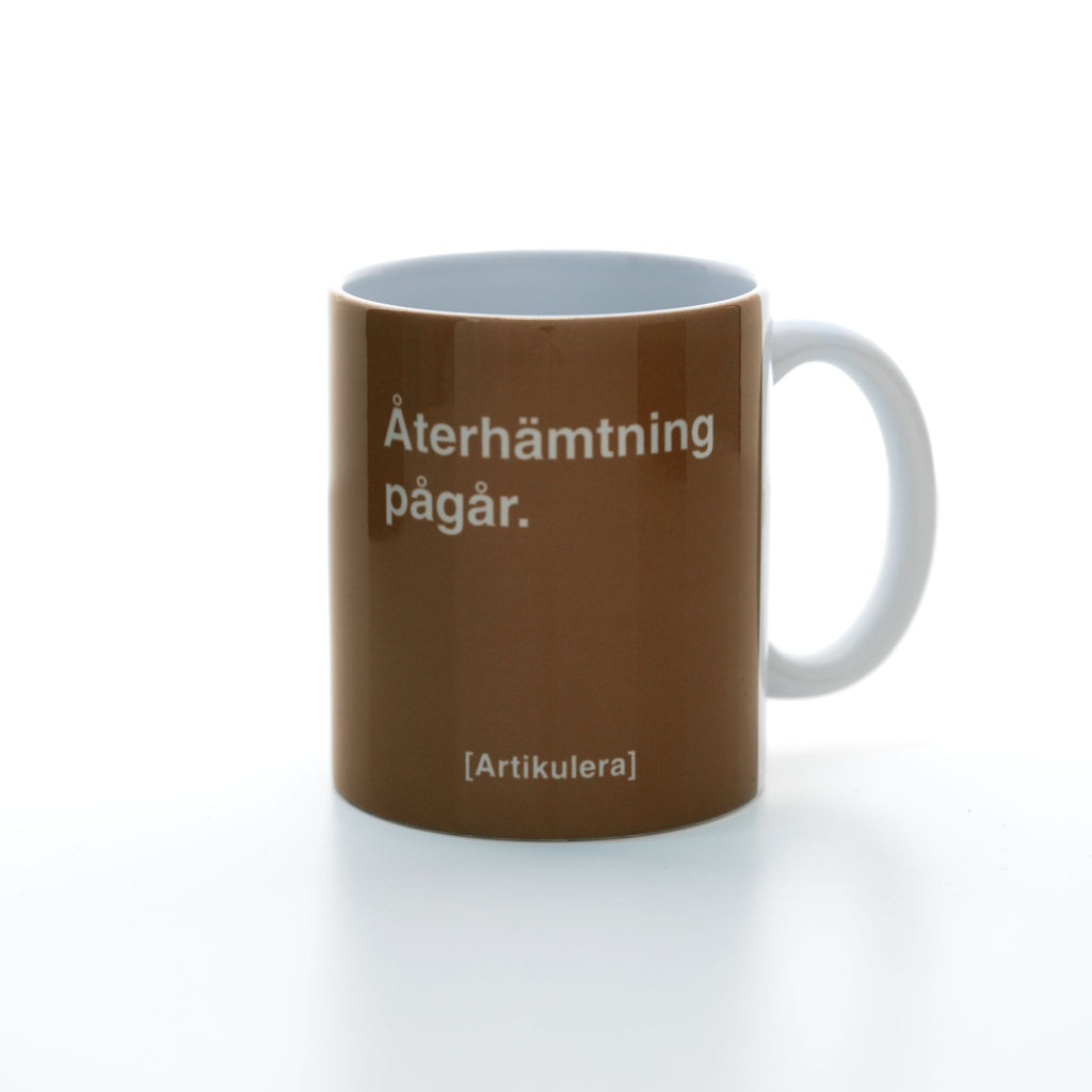 Vit mugg med mörkbrunt tryck, vit text ”Återhämtning pågår.” samt vit Artikulera-logotyp