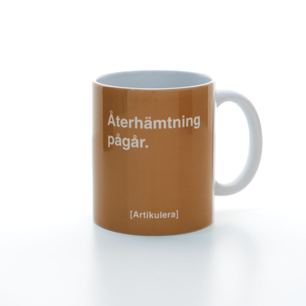 Vit mugg med mörk orangefärgat tryck, vit text ”Återhämtning pågår.” samt vit Artikulera-logotyp