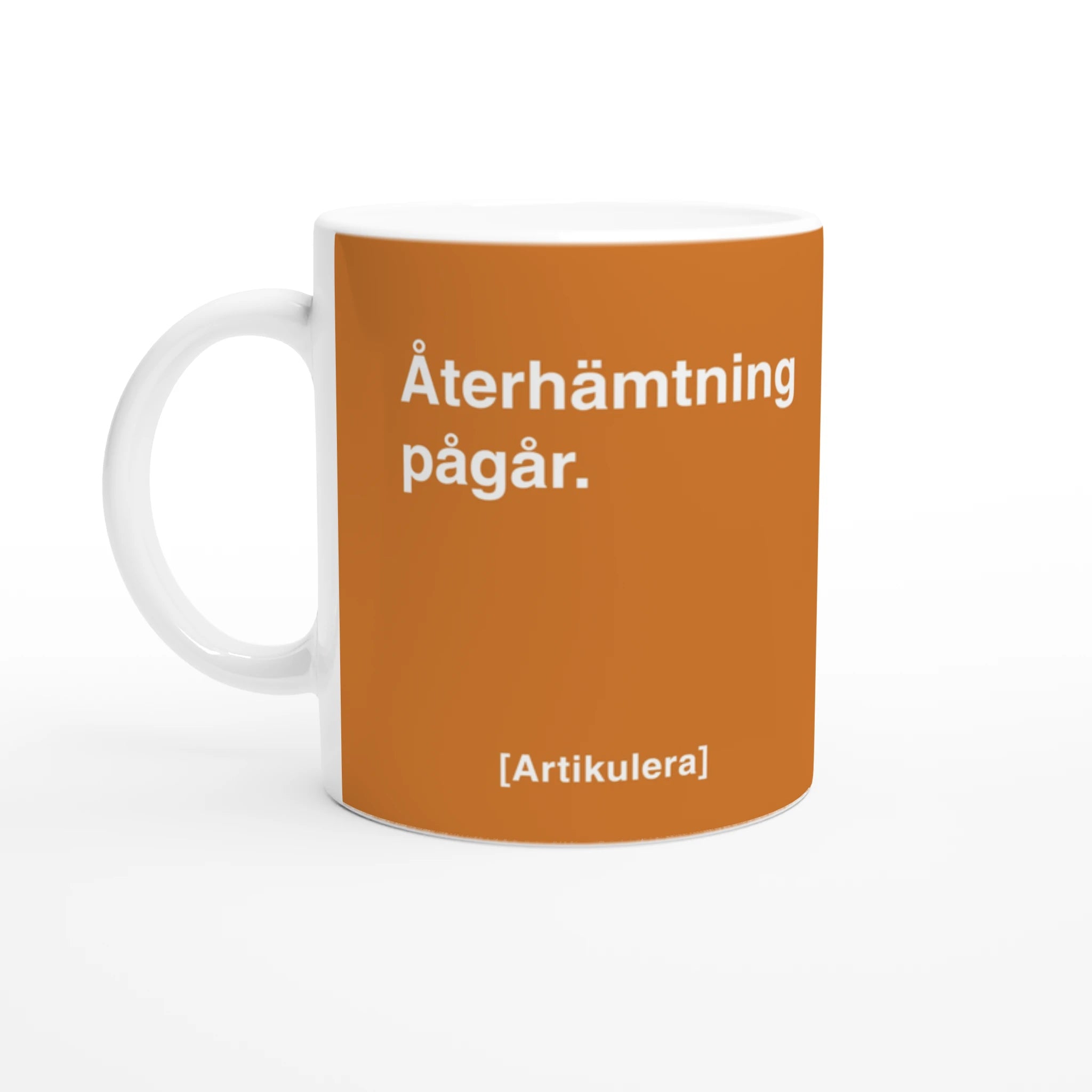 Vit mugg med mörk orangefärgat tryck, vit text ”Återhämtning pågår.” samt vit Artikulera-logotyp