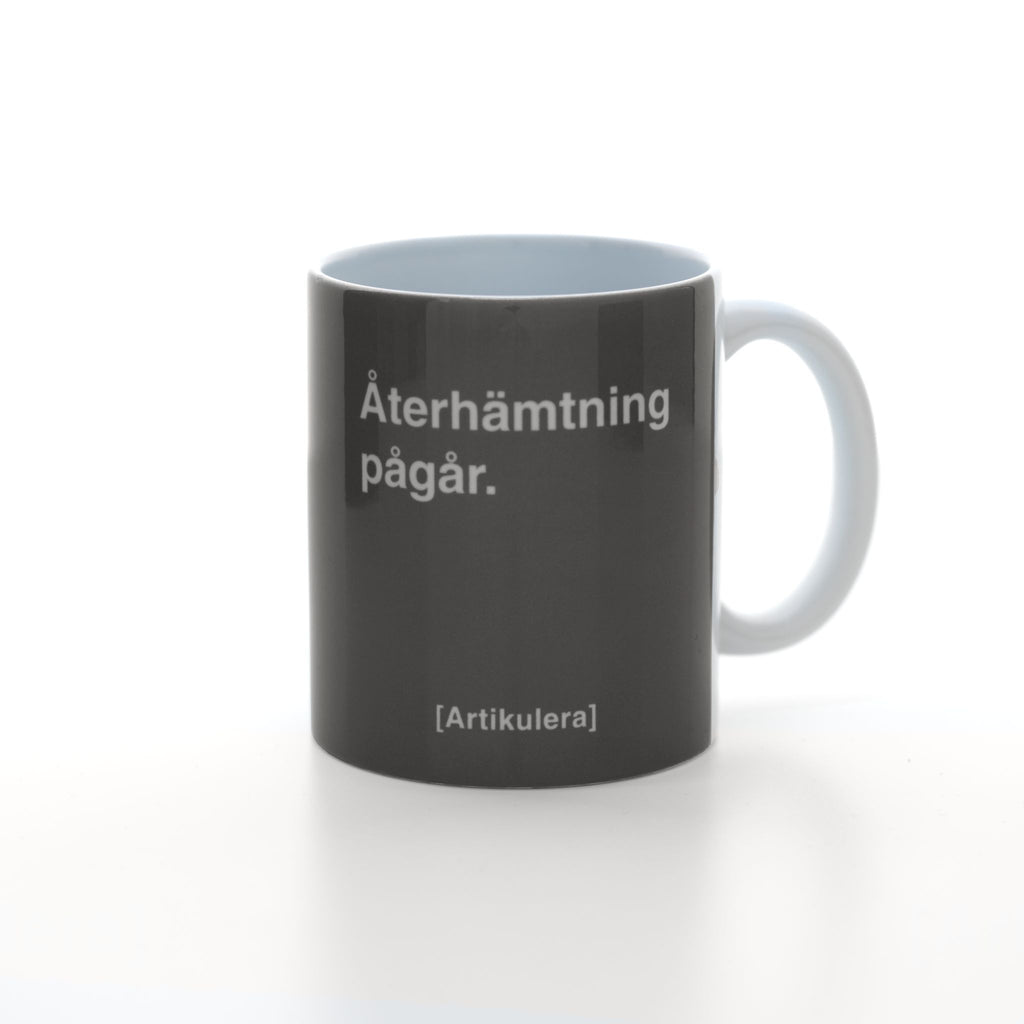 Vit mugg med mörkt rockigt grått tryck, vit text ”Återhämtning pågår.” samt vit Artikulera-logotyp