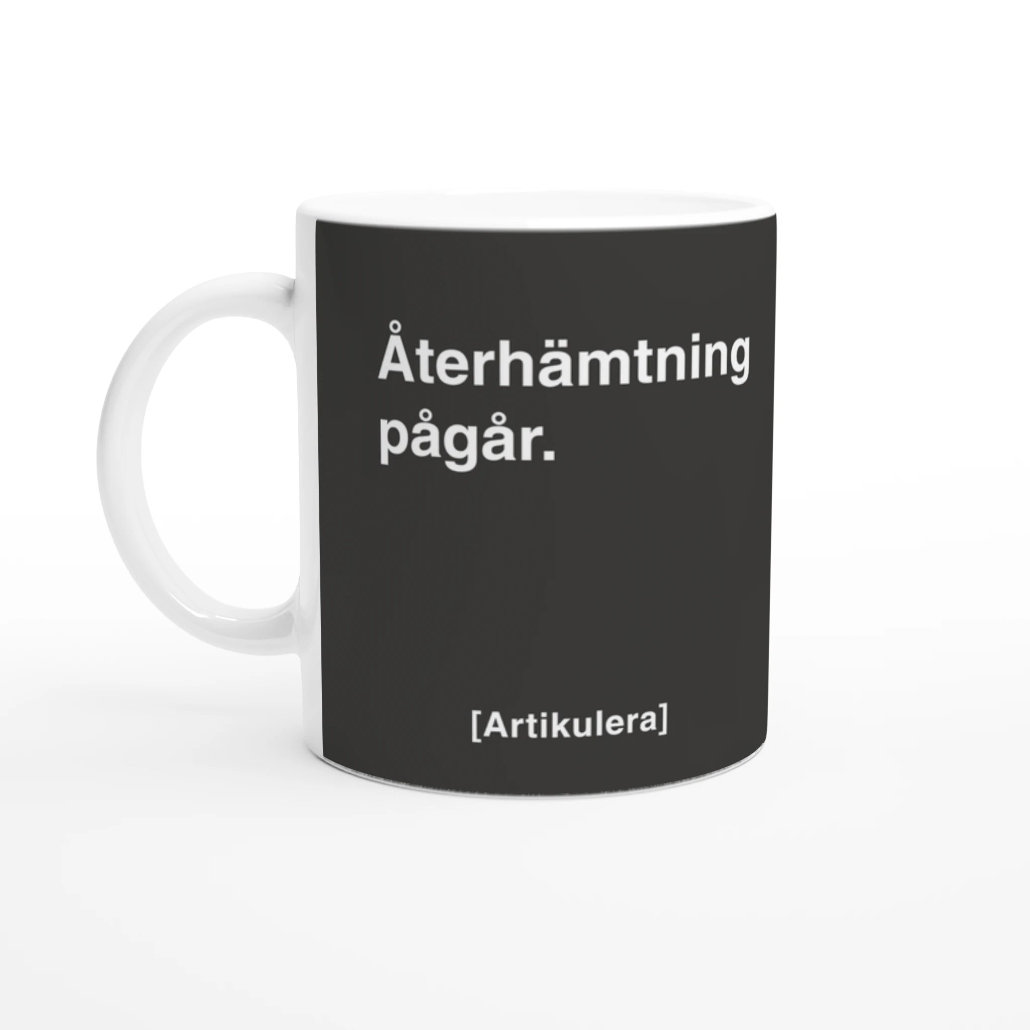Vit mugg med mörkt rockigt grått tryck, vit text ”Återhämtning pågår.” samt vit Artikulera-logotyp