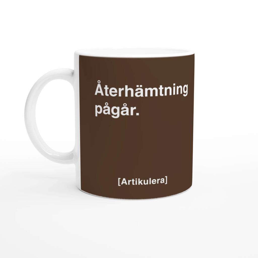 Vit mugg med mörkbrunt tryck, vit text ”Återhämtning pågår.” samt vit Artikulera-logotyp