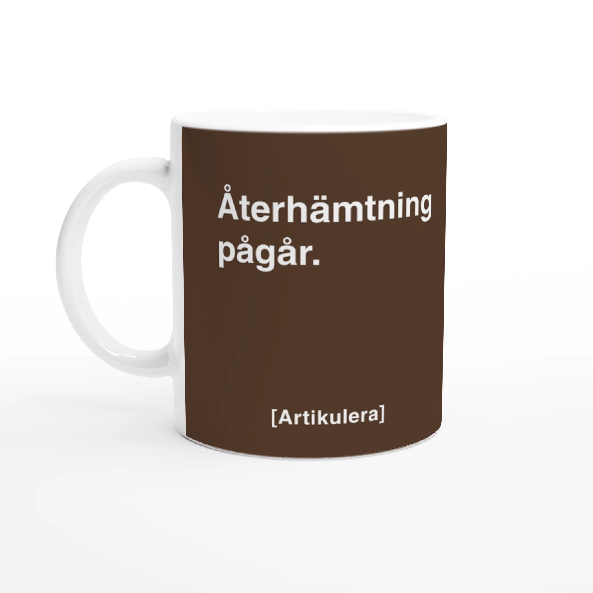 Vit mugg med mörkbrunt tryck, vit text ”Återhämtning pågår.” samt vit Artikulera-logotyp