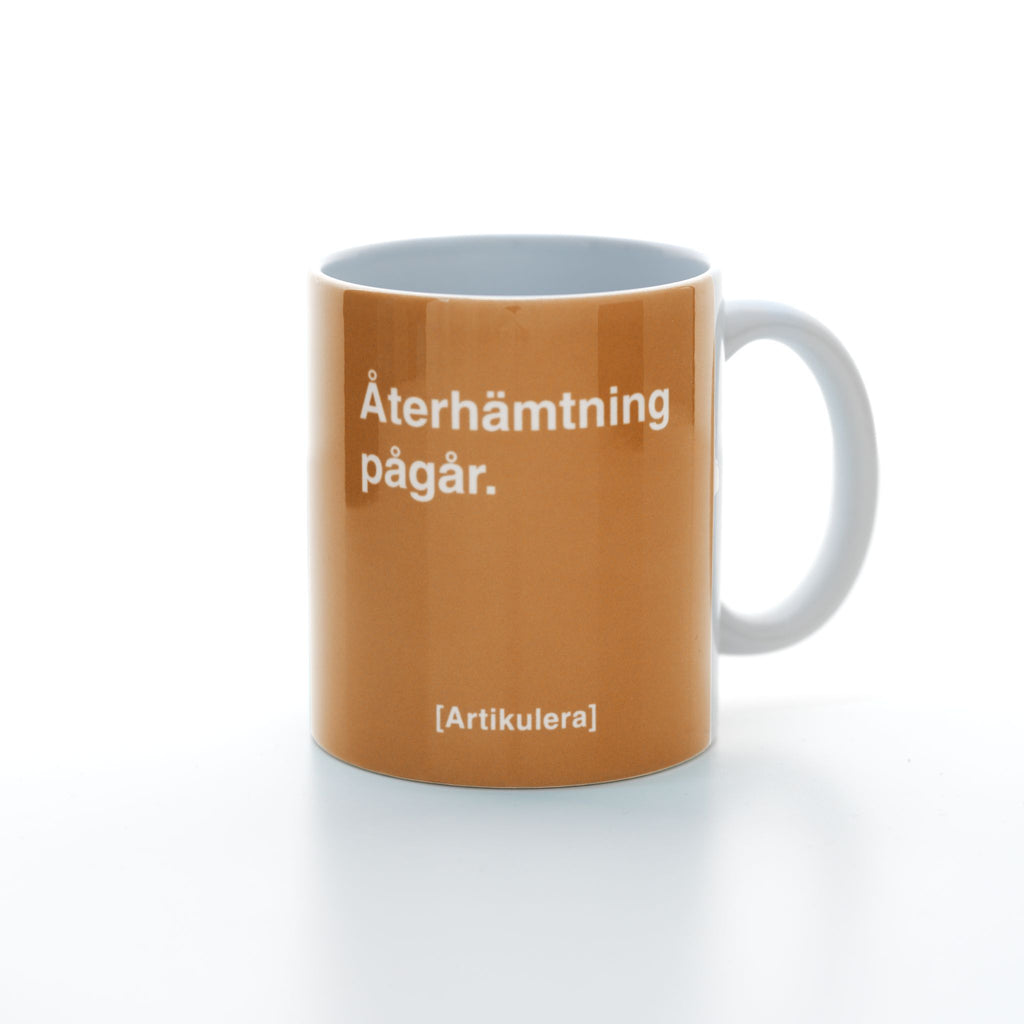 Vit mugg med orangefärgat tryck, vit text ”Återhämtning pågår.” samt vit Artikulera-logotyp
