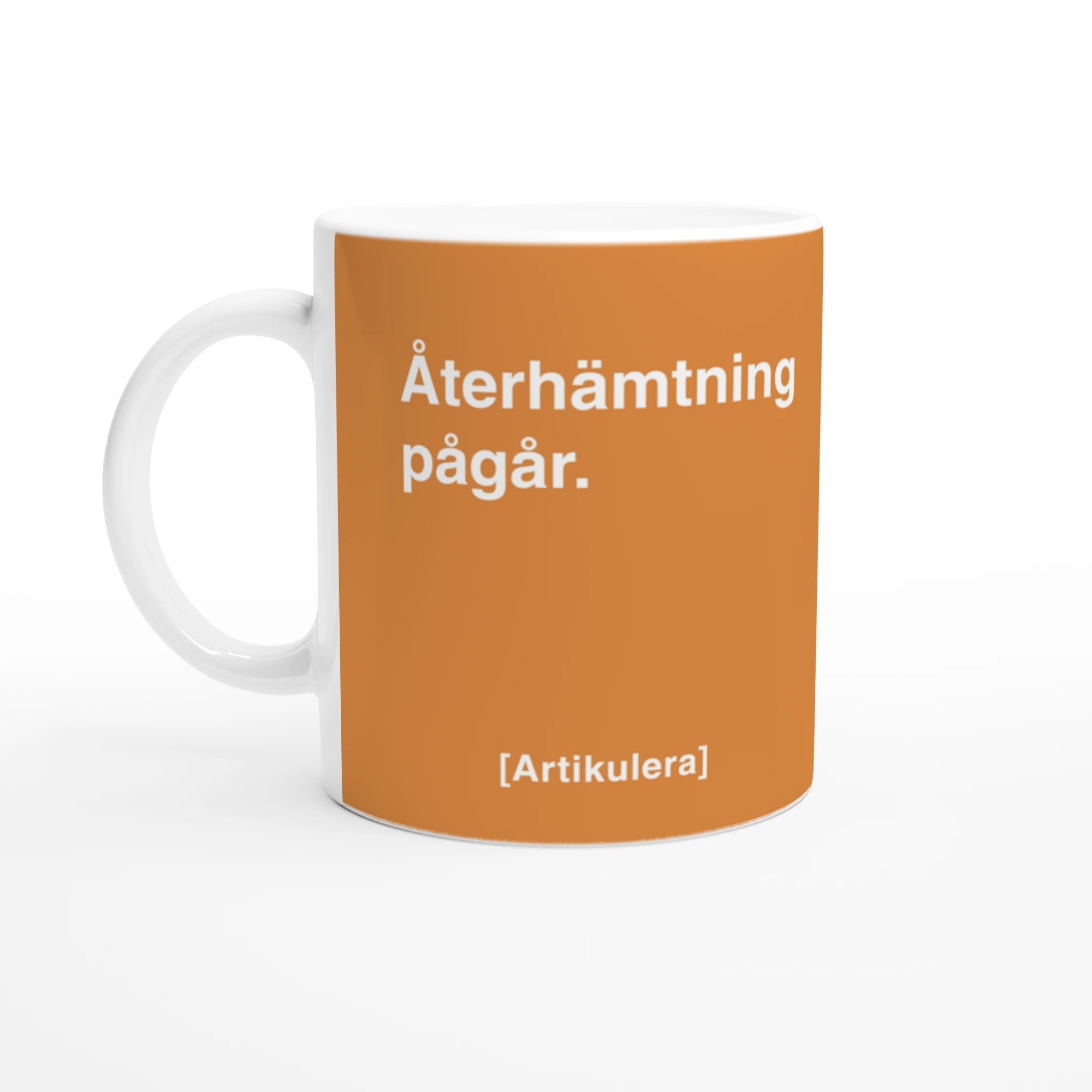 Vit mugg med orangefärgat tryck, vit text ”Återhämtning pågår.” samt vit Artikulera-logotyp