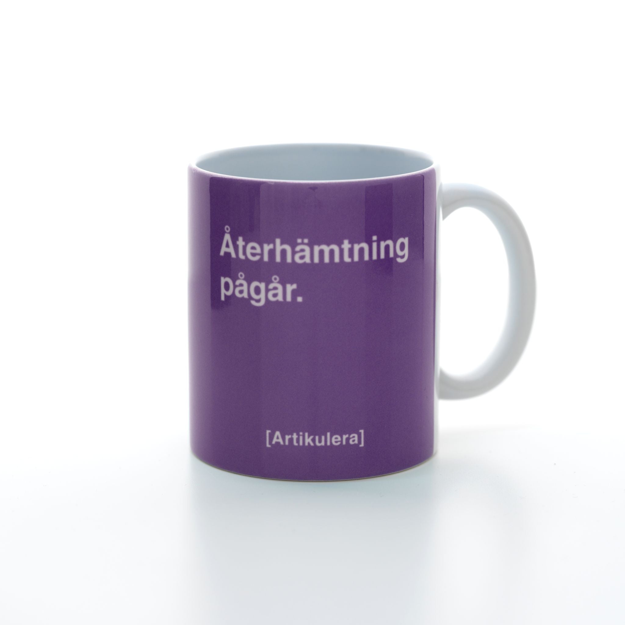 Vit mugg med plommonlila tryck, vit text ”Återhämtning pågår.” samt vit Artikulera-logotyp