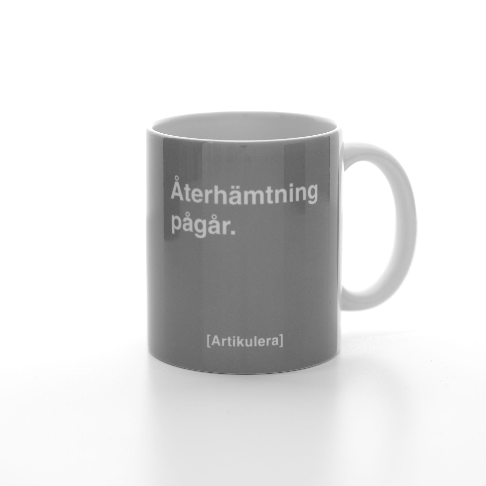 Vit mugg med rockigt grått tryck, vit text ”Återhämtning pågår.” samt vit Artikulera-logotyp