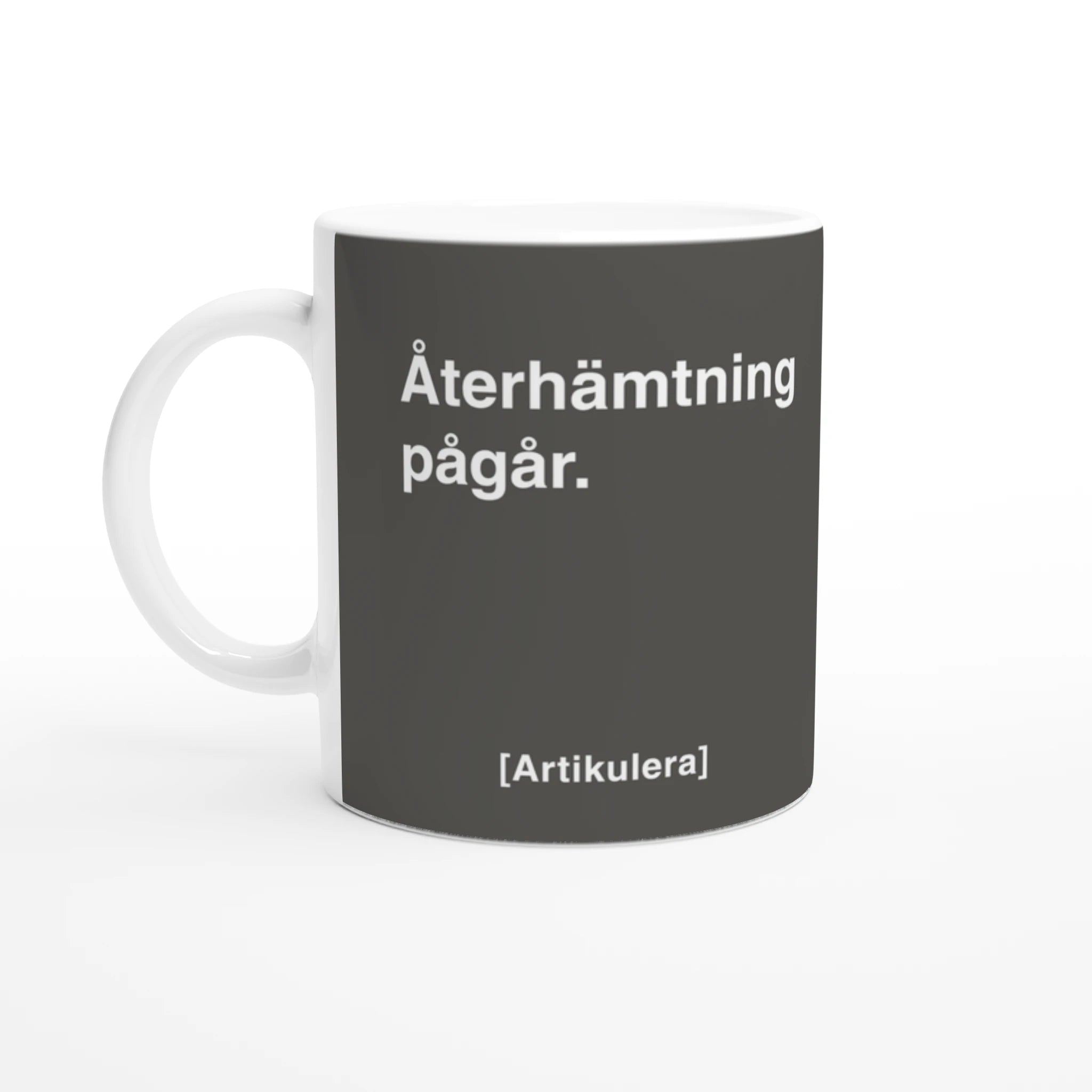 Vit mugg med rockigt grått tryck, vit text ”Återhämtning pågår.” samt vit Artikulera-logotyp