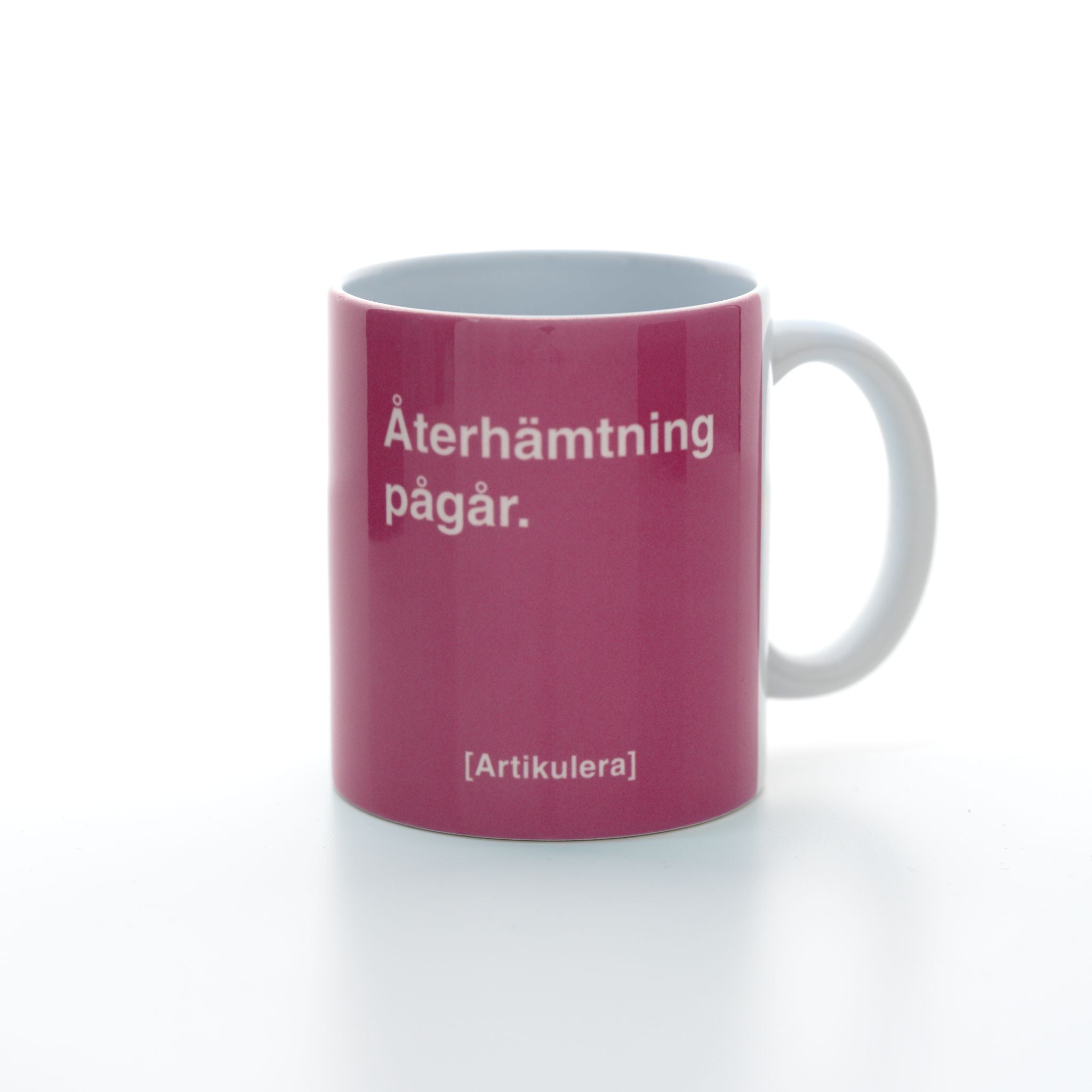 Vit mugg med rosa tryck, vit text ”Återhämtning pågår.” samt vit Artikulera-logotyp