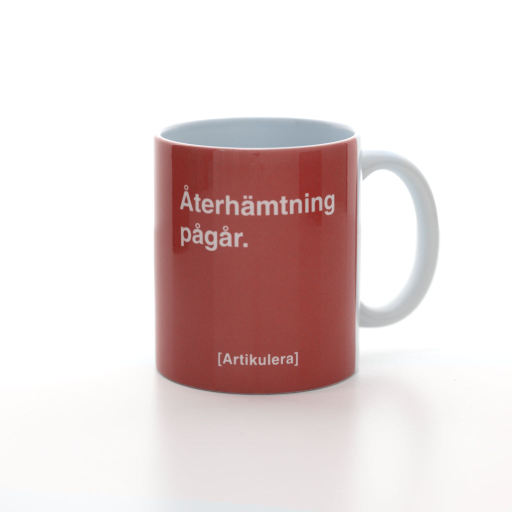 Vit mugg med tomatrött tryck, vit text ”Återhämtning pågår.” samt vit Artikulera-logotyp