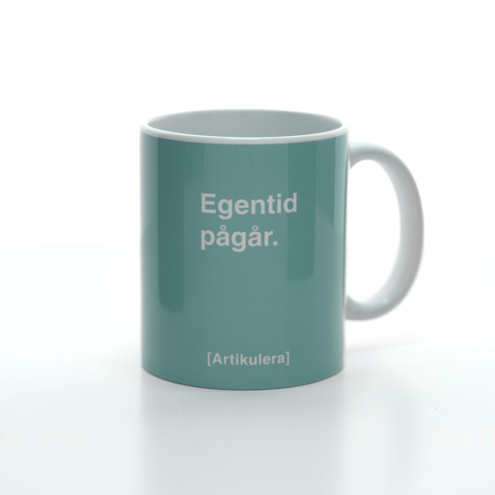 Vit mugg med ljust blågrönt tryck, vit text ”Egentid pågår.” samt vit Artikulera-logotyp