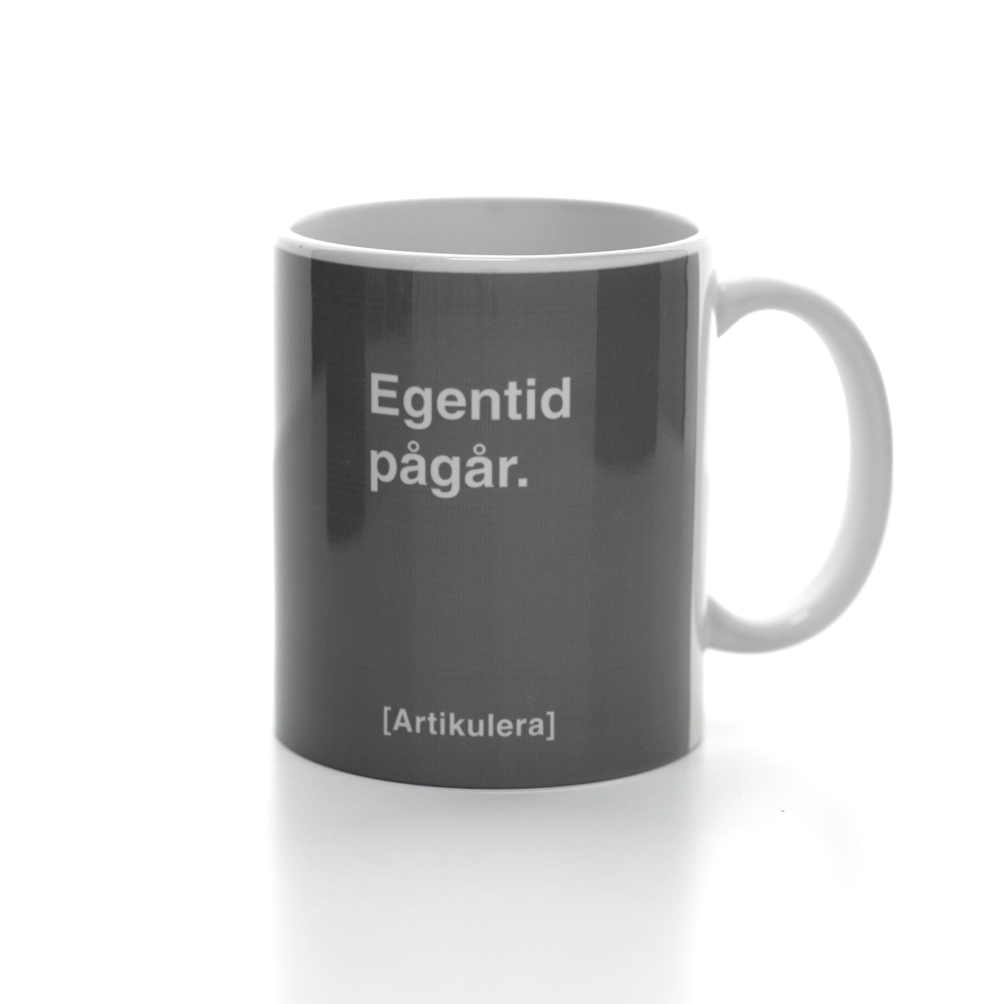 Vit mugg med mörkt rockigt grått tryck, vit text ”Egentid pågår.” samt vit Artikulera-logotyp