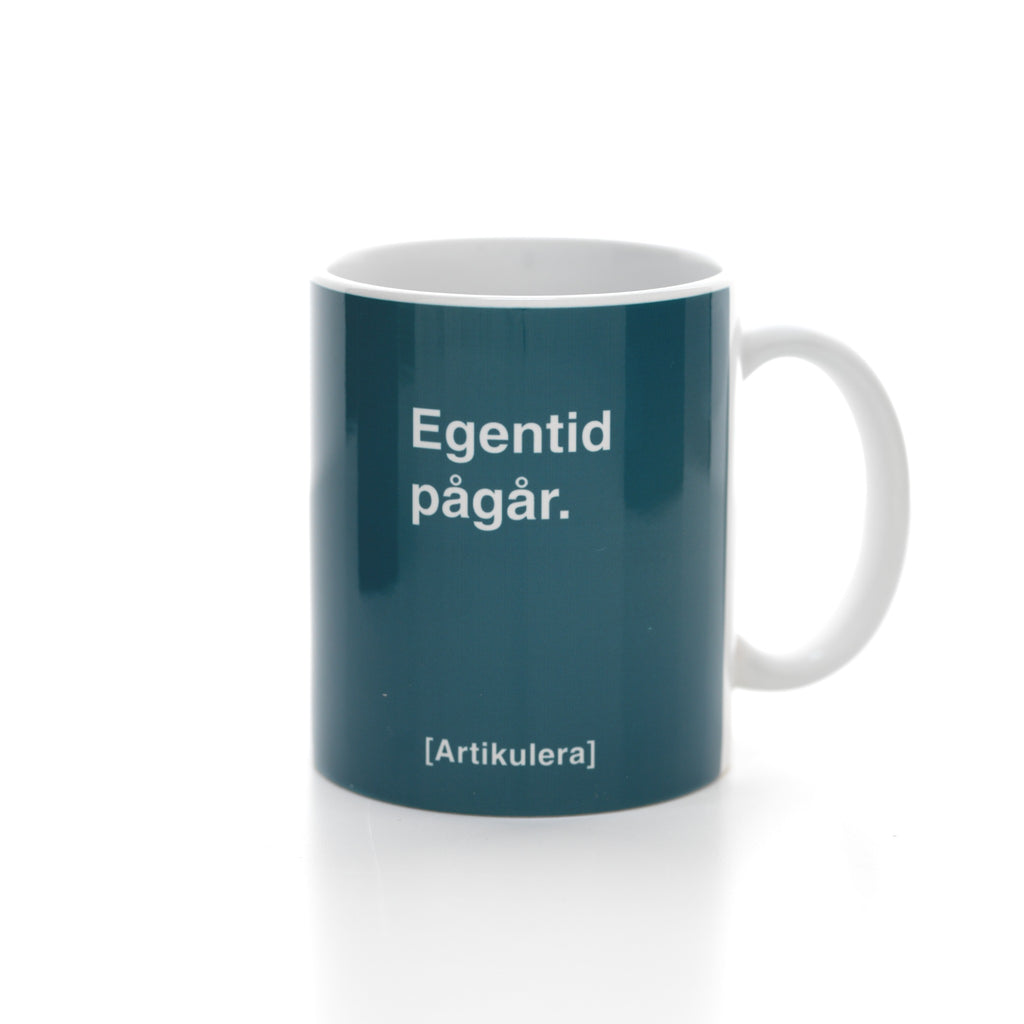 Vit mugg med mörk turkosgrönt tryck, vit text ”Egentid pågår.” samt vit Artikulera-logotyp