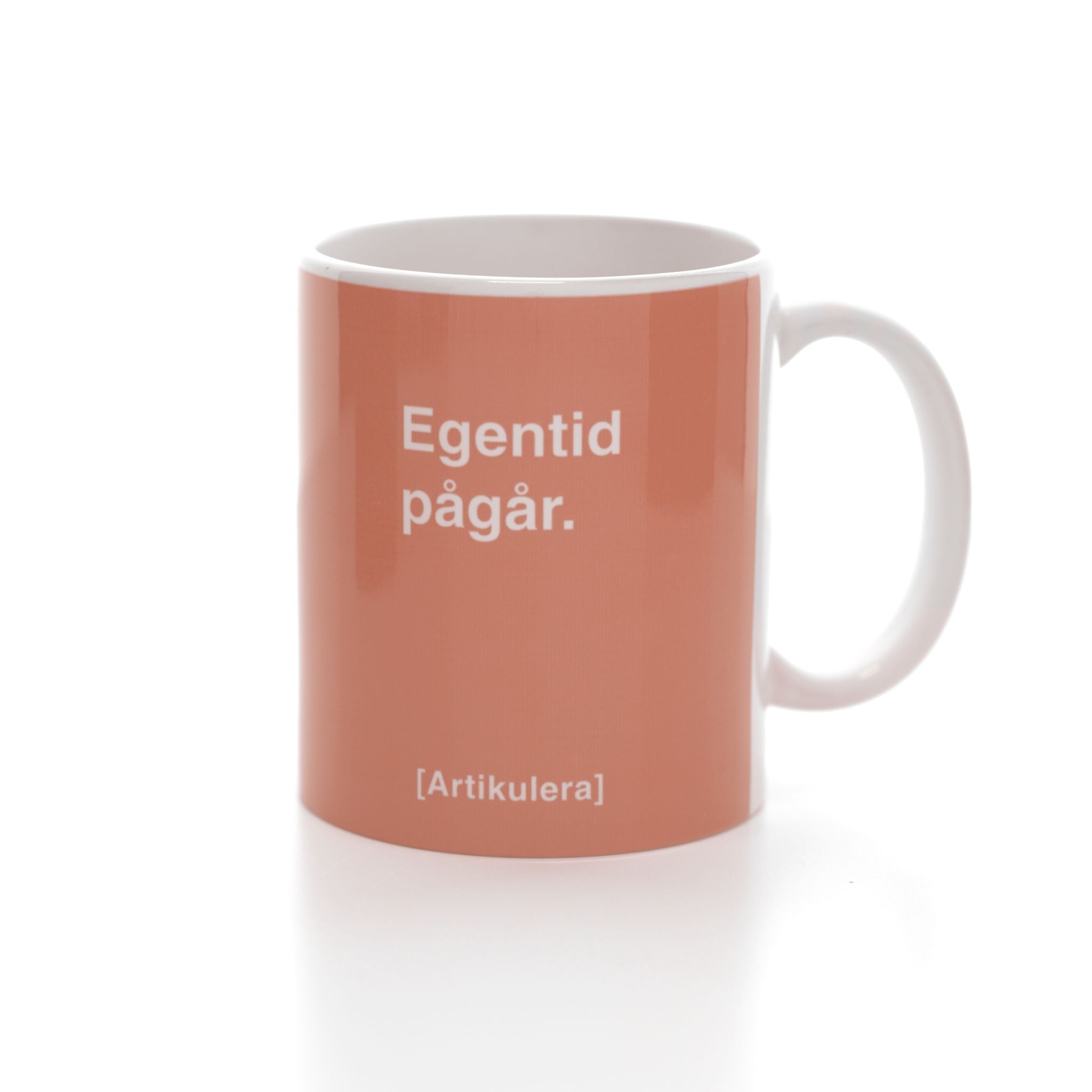 Vit mugg med persikofärgat tryck, vit text ”Egentid pågår.” samt vit Artikulera-logotyp