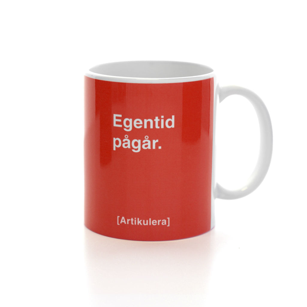 Vit mugg med tomatrött tryck, vit text ”Egentid pågår.” samt vit Artikulera-logotyp