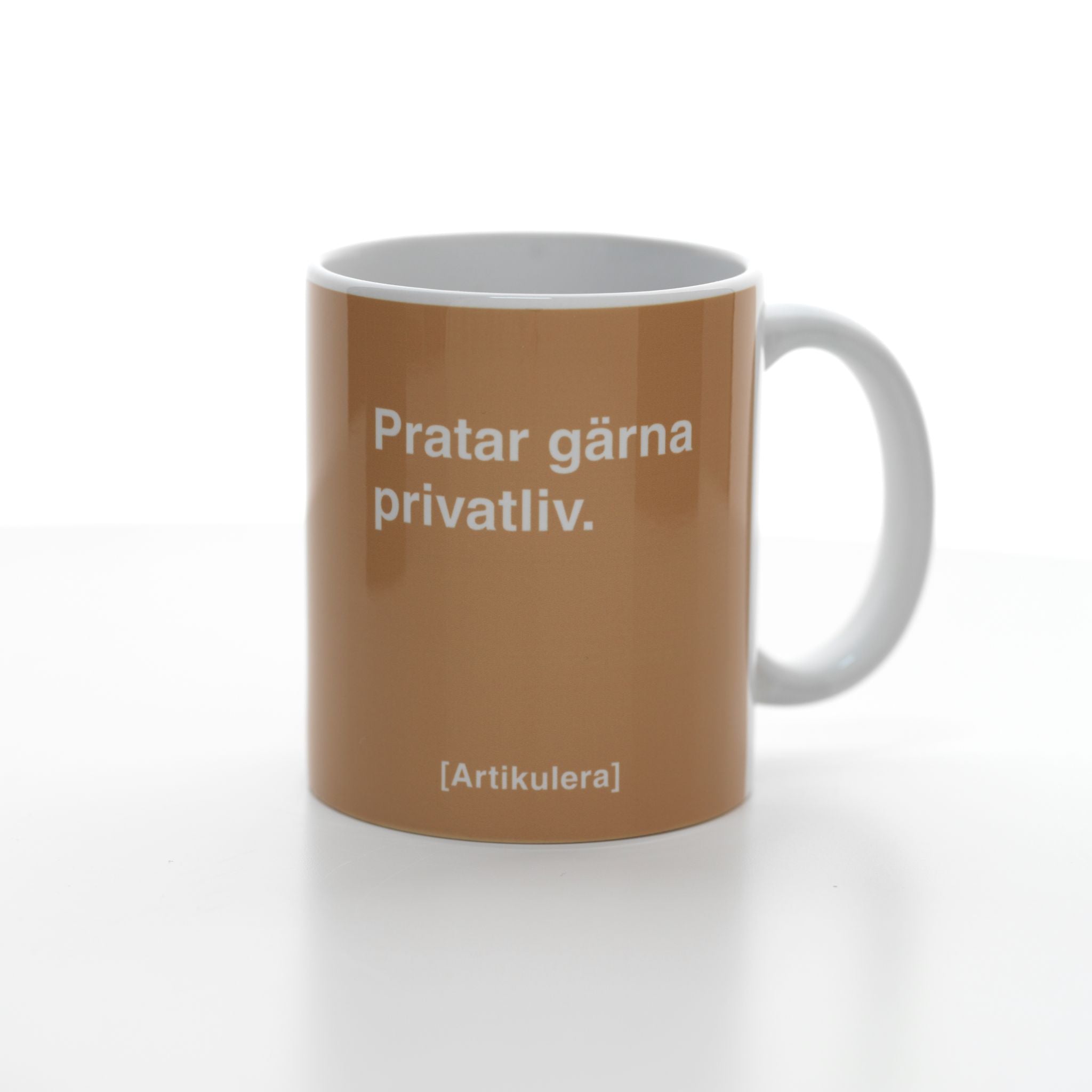 Vit mugg med mediumbrun tryck, vit text ”Pratar gärna privatliv.” samt vit Artikulera-logotyp