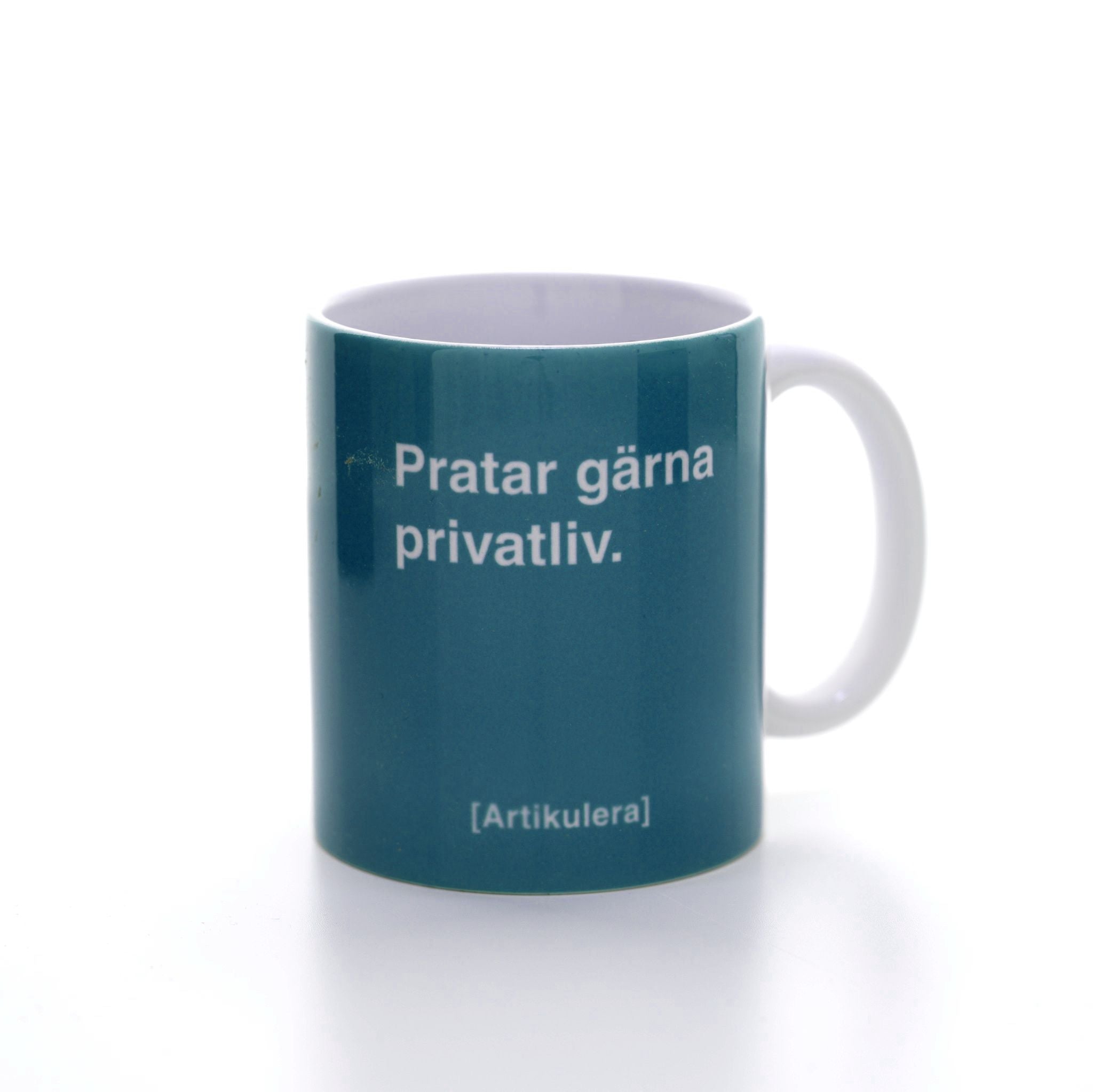 Vit mugg med mörkt turkosgrönt tryck, vit text ”Pratar gärna privatliv.” samt vit Artikulera-logotyp
