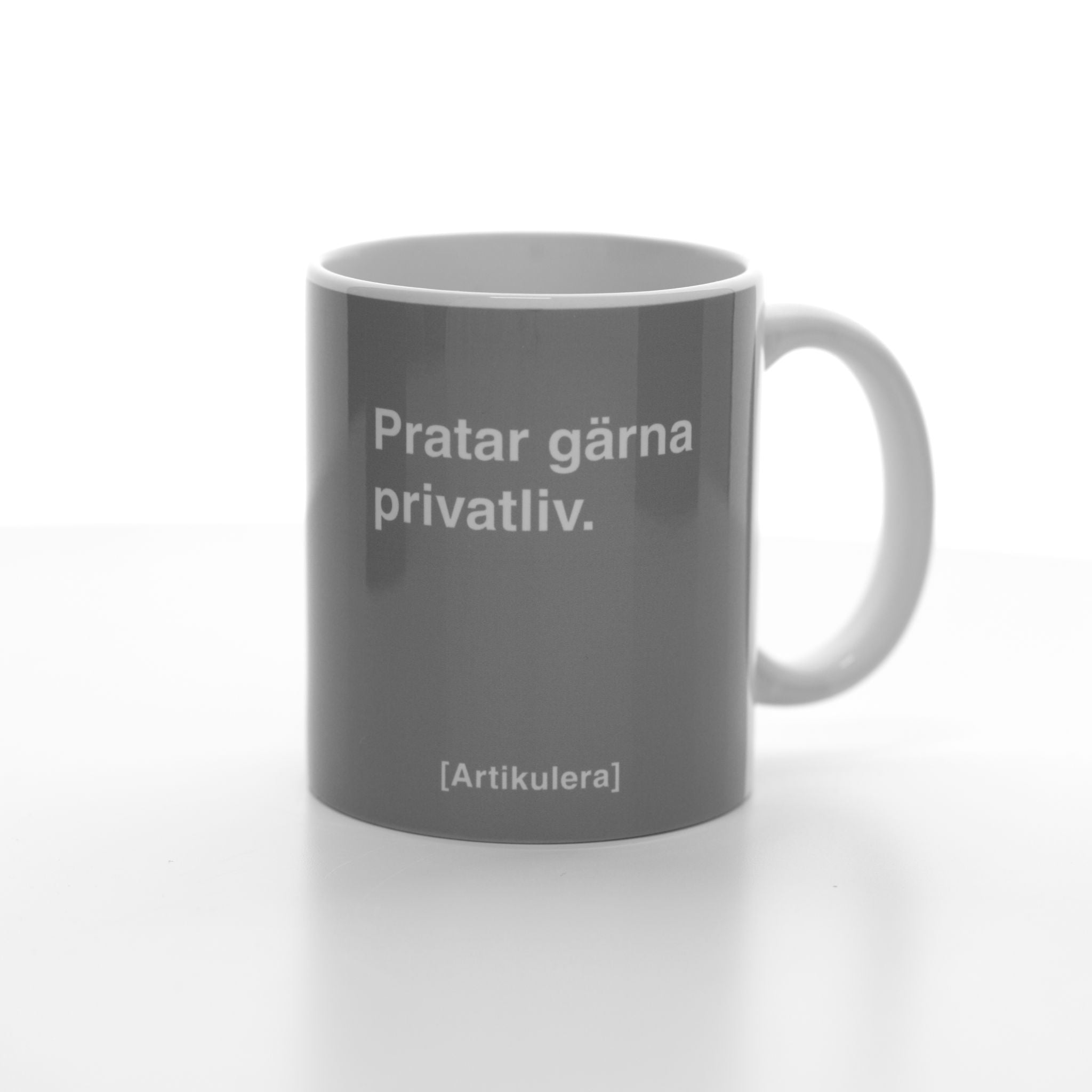 Vit mugg med mörkt rockigt grått tryck, vit text ”Pratar gärna privatliv.” samt vit Artikulera-logotyp